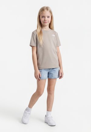 Jeune fille debout, portant un t-shirt taupe à manches courtes, un short en jean bleu clair, des chaussettes blanches et des baskets blanches sur un fond uni.