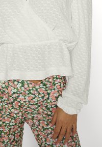 T-shirt branca e texturizada com mangas longas e punhos franzidos, combinada com calças com padrão floral em tons de rosa e verde.