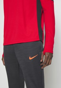 Rode atletische shirt met lange mouwen en zwarte zijpanelen; gecombineerd met donkergrijze gebreide broek met een oranje Nike-logo op de linkerbovenbeen.