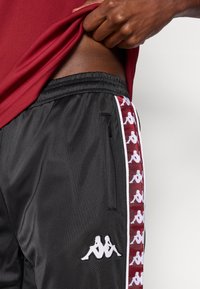 Czarne sportowe spodnie z czerwonym bocznym panelem ozdobionym białymi logo Kappa. Talia jest elastyczna, a po boku znajduje się kieszeń na suwak.