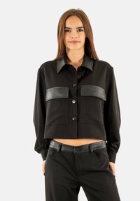 Chaqueta negra corta con cuello, presenta detalles de cuero en los bolsillos del pecho y los hombros, y cierre de botones en la parte delantera.