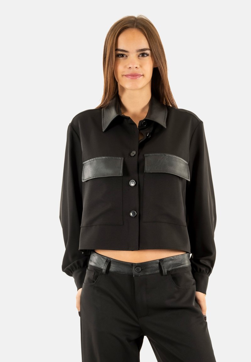 Chaqueta negra corta con cuello, presenta detalles de cuero en los bolsillos del pecho y los hombros, y cierre de botones en la parte delantera.