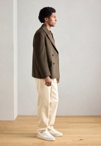 Olijfgroene double-breasted wollen blazer, roomkleurige wijde broek en witte sneakers. De gladde textuur en contrasterende kleuren benadrukken het geheel.
