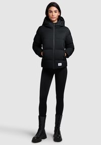 Sort polstret jakke med hætte, med en frontlynlås, sidelommer og et logo-patch. Båret med sorte leggings og snørebåndsstøvler.