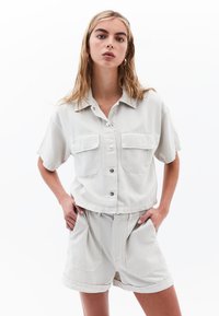 Jasnoszara koszula krótki rękaw z dwiema kieszeniami z przodu i zapięciem na guziki, zestawiona z luźnymi szortami w tym samym kolorze. Gładki materiał bawełniany.