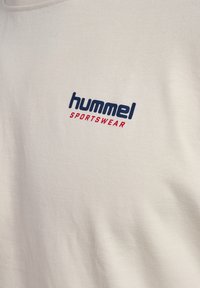 Tessuto color crema con scritte blu "hummel" e rosse "SPORTSWEAR" stampate, che indicano il logo di un marchio di abbigliamento sportivo.