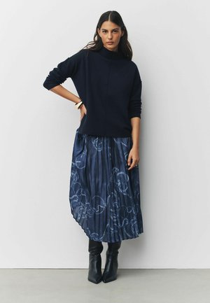 Marineblauer Rollkragenpullover mit langen Ärmeln, kombiniert mit einem marineblauen Faltenrock mit dezentem Blumenmuster und schwarzen Stiefeln mit Absatz.