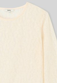 Top en dentelle crème avec un col rond et des manches longues. Le tissu présente un motif floral et une apparence texturée. Taille S.