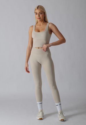 Mujer con cabello largo y rubio, vistiendo un top corto sin mangas beige y leggings a juego, calcetines blancos y zapatillas blancas, de pie sobre un fondo gris.