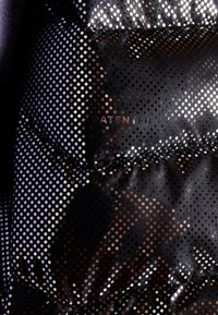 Close-up van zwart doorgestikt stof met kleine ronde metalen stippen en gestikte naden, met vaag zichtbare tekst "PATENTED".