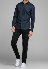 Man som bär en marinblå skjorta med knappar, svarta byxor, vita sneakers och en silverring på sin vänstra hand, stående mot en grå bakgrund.