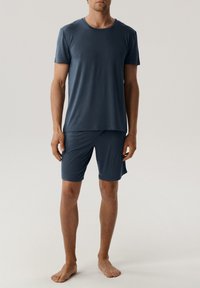 Mężczyzna stojący boso, ubrany w gładką, granatową koszulkę z krótkim rękawem i pasujące szorty do kolan na jasnym tle.