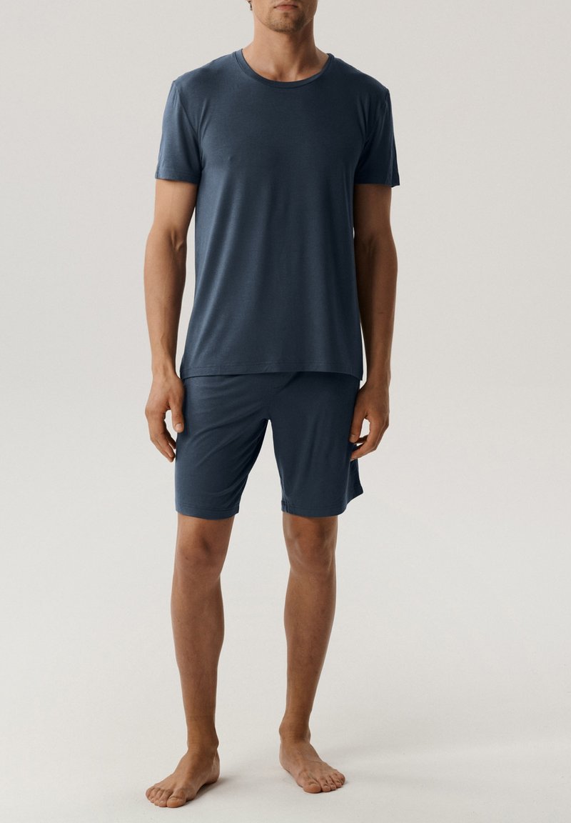 Mężczyzna stojący boso, ubrany w gładką, granatową koszulkę z krótkim rękawem i pasujące szorty do kolan na jasnym tle.