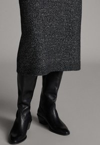 Schwarze kniehohe Stiefel aus Leder mit spitz zulaufender Spitze, Blockabsatz und dekorativen Nähten, kombiniert mit einem strukturierten, dunkelgrauen Strickkleid.