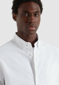 Giovane uomo dalla pelle scura che indossa una camicia bianca con colletto abbottonata su uno sfondo chiaro e semplice.