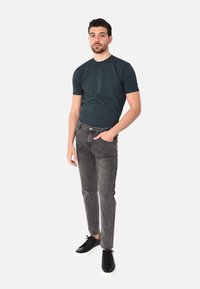 T-shirt grigia, jeans slim-fit grigi e sneakers nere con suole bianche. Il modello sta in piedi con una mano in tasca contro uno sfondo bianco.