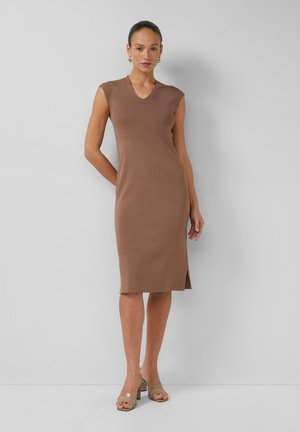 Kobieta w brązowej sukience bez rękawów, sięgającej kolan z bocznym rozcięciem, nosząca złote kolczyki-koła i beżowe sandały na obcasie, stojąca na tle szarej ściany.