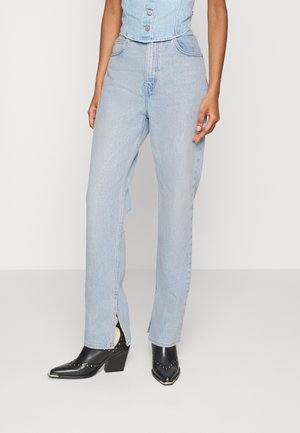 Personne portant un jean taille haute bleu clair avec des fentes à l'avant et des bottines noires à bout pointu cloutées.