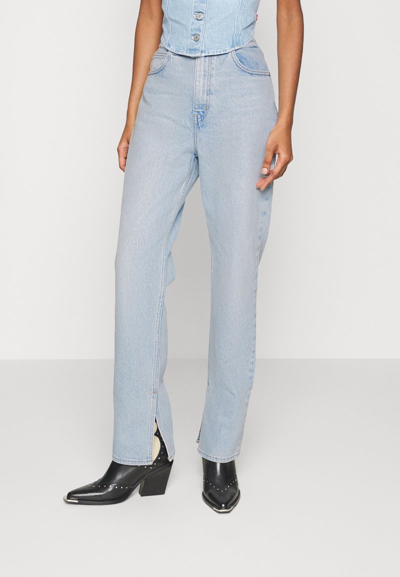 Personne portant un jean taille haute bleu clair avec des fentes à l'avant et des bottines noires à bout pointu cloutées.