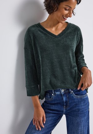 Haut à manches longues texturé vert foncé avec un col en V, associé à un jean en denim bleu. Le haut a des manches larges, une coupe décontractée et un motif subtil.