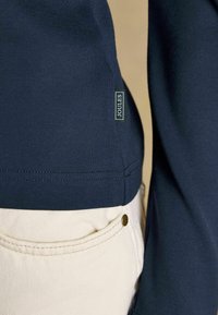 Temno modra bombažna majica s kratkimi rokavi, z oznako na robu. Nosite jo z svetlobezžimi kavbojkami, ki prikazujejo podrobnosti žepa.