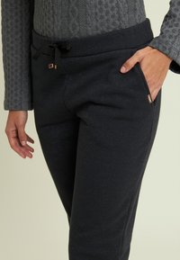 Persona che indossa pantaloni jogger neri con vita regolabile e maglia grigia a maniche lunghe con texture, con una mano in tasca, su uno sfondo chiaro.