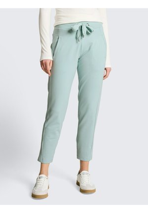 Person trägt hellblaue knöchellange Hose mit vorderer Schleife und weiße Turnschuhe, steht vor einem schlichten Hintergrund.