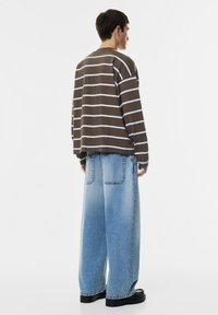 Persoon met de rug naar de camera draagt een bruine en blauwe gestreepte lange mouwen shirt, losse lichtblauwe spijkerbroek en zwarte schoenen op een witte achtergrond.