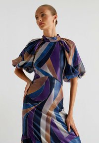 Sukienka wykonana z jedwabistego materiału z odważnymi geometrycznymi wzorami w kolorach granatowym, purpurowym i miedzianym. Bufiaste rękawy i projekt z wysokim dekoltem.
