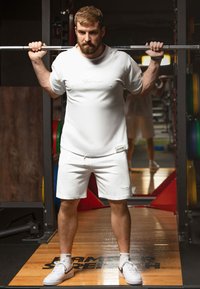 Witte sportieve t-shirt en shorts, met logo-details. Het model heft een halter en staat op een gymvloer met apparatuur op de achtergrond.