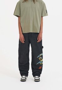 Olive green short-sleeve t-shirt paired with black cargo pants featuring colorful dragon embroidery, complemented by black and white sneakers.