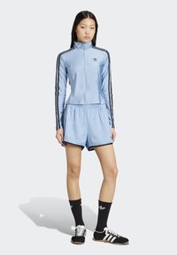 Ung kvinna som står klädd i en ljusblå Adidas träningsjacka och shorts med svarta ränder, svarta Adidas-strumpor och svarta sneakers.