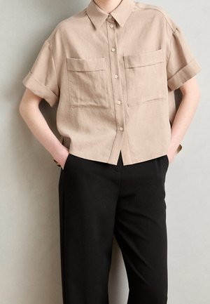 Beige linnen shirt met korte mouwen, knoopsluiting en twee voorzakken. Draagt bij zwarte broek. Gladde textuur en ontspannen pasvorm.