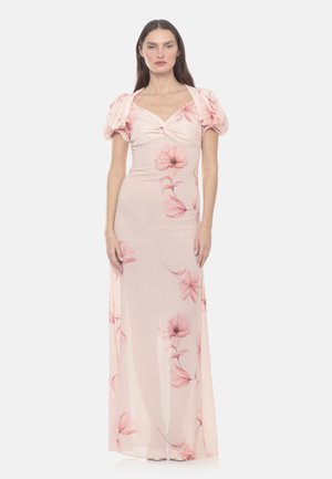 Donna che indossa un abito lungo rosa pallido con maniche a sbuffo e grandi stampe floreali rosa morbide, in piedi davanti a uno sfondo semplice.