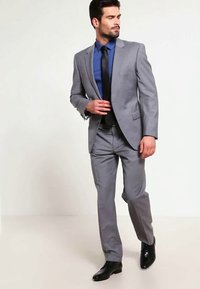 Άνδρας με γκρι κοστούμι, μπλε πουκάμισο και μαύρη γραβάτα περπατά με το χέρι κοντά στο κουμπί του σακακιού, κοιτάζοντας προς τα αριστερά.