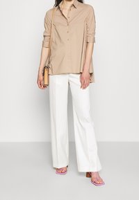 Camicia beige con maniche arrotolate, abbinata a pantaloni bianchi a gamba larga. Gli accessori includono una piccola borsa beige e sandali rosa.