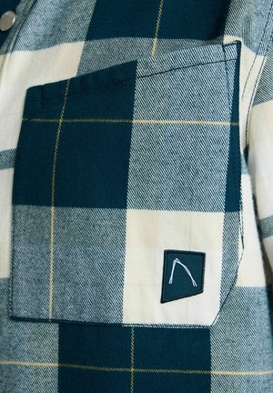 Nærbillede af et blå- og hvidternet stof med en firkantet lomme, der har en lille sort lap med et hvidt abstrakt mønster.