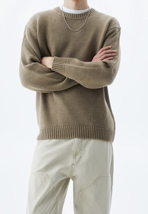 Person trägt einen braunen Strickpullover, ein weißes Schichthemd, eine silberne Kettenhalskette und hellbeige Hose, steht mit verschränkten Armen.