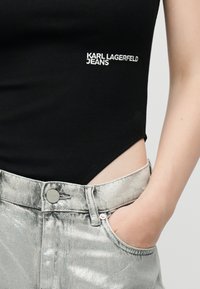 Κοντινό πλάνο ενός ατόμου που φοράει μαύρο τοπ με το κείμενο «KARL LAGERFELD JEANS» και ασημί μεταλλικά τζιν με το ένα χέρι στην τσέπη.