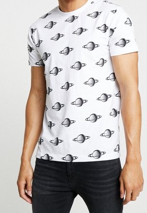 Weißes Kurzarm-T-Shirt mit wiederholtem schwarz-weißem Planeten-mit-Ringen-Muster, getragen zu schwarzen Jeans von einer Person, die vom Hals bis zur Hüfte gezeigt wird.