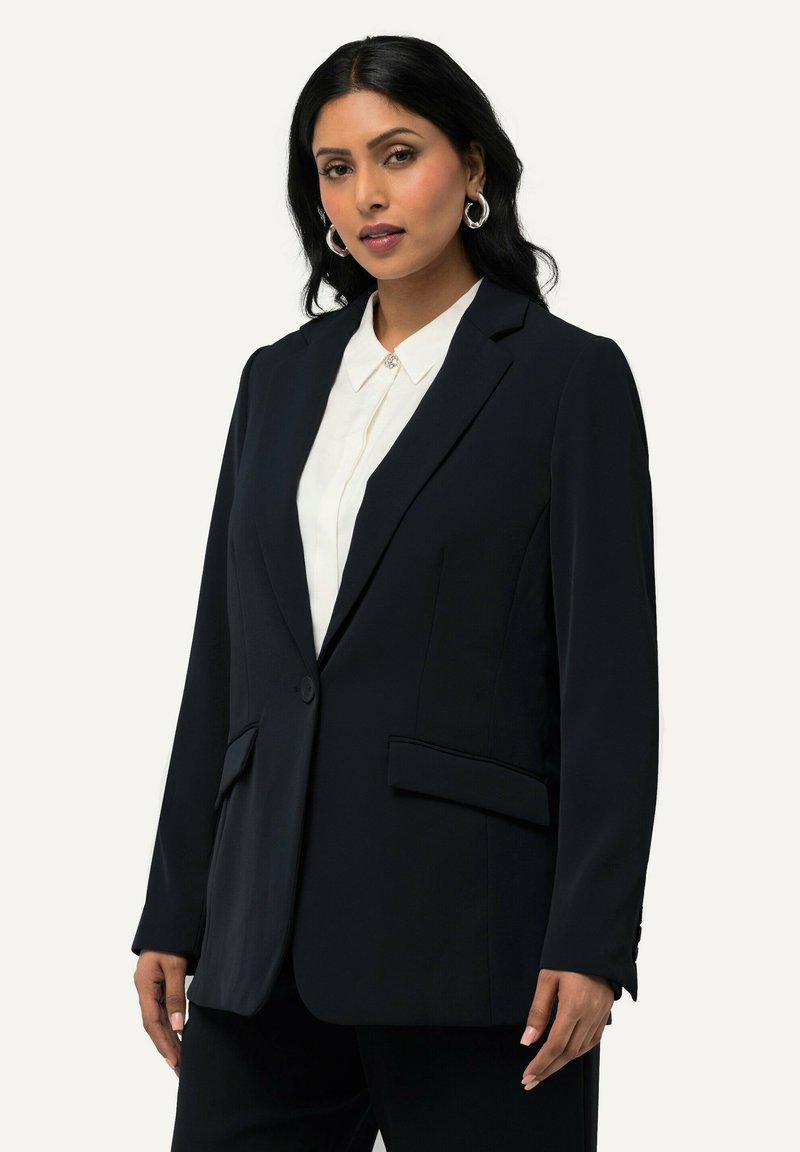 Blazer bleu marine en tissu lisse avec une fermeture à un bouton, deux poches avant et coupe ajustée ; porté sur une chemise blanche à col.