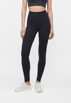 Kvinde iført sorte højtaljede leggings, sort crop top og hvide sneakers står mod en ensfarvet hvid baggrund.