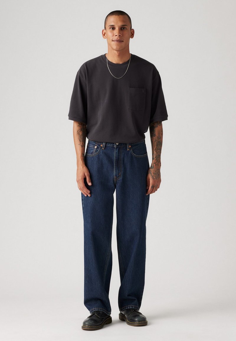 Schwarzes T-Shirt mit kurzen Ärmeln und einer Brusttasche, kombiniert mit locker sitzenden blauen Jeans. Schwarze Schuhe runden das Outfit ab.