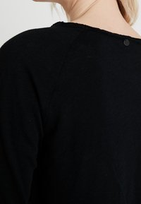 Svart tröja med rund halsringning, texturerad tyg och en liten knappdetalj i nacken.