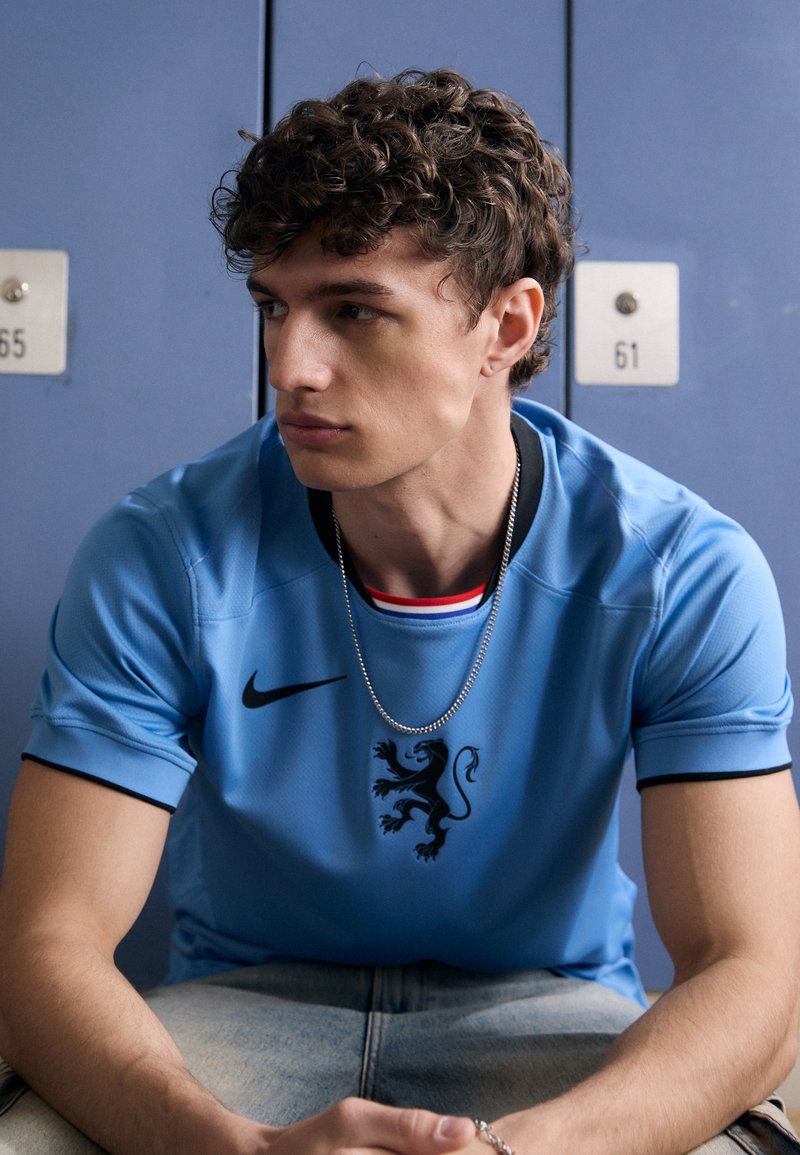 Maglia sportiva Nike di colore azzurro chiaro con un emblema di leone nero e bordature nere a contrasto, abbinata a pantaloni in denim chiaro. Collana di catena argentata visibile.