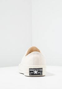 Sneaker beige in tela a basso con tomaia testurizzata e suola in gomma, dotato di un'etichetta nera con testo bianco e dettagli a forma di stella sul tallone.