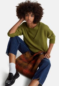 Olivově zelený žebrovaný svetr s výstřihem do V, spárovaný s modrými džíny s třepenými lemy. Černé mokasíny a vzorovaná taška v teplých tónech.