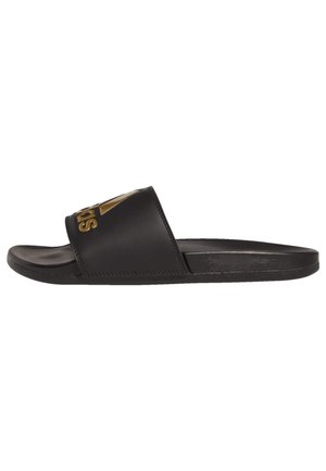 Sandalias de slide negras hechas de material sintético suave con una correa ancha que presenta el nombre de la marca en dorado. Suela plana con textura mínima.