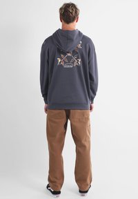 Sweat à capuche gris avec un motif floral et le texte "OXBOW" dans le dos, associé à un pantalon cargo marron et des baskets noires.