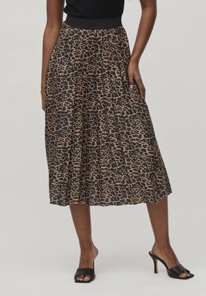 Kobieta nosząca plisowaną spódnicę midi w brązowo-czarny wzór w cętki, czarną bluzkę oraz czarne mulesy na obcasie, stojąca na neutralnym tle.